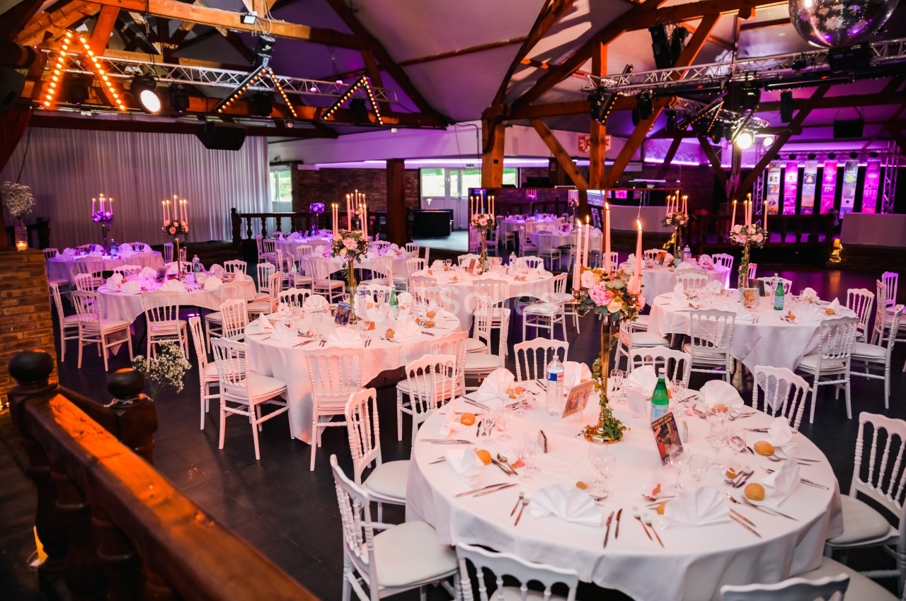 Salle de réception décorée avec des tables rondes dressées, chandeliers et éclairage tamisé dans une ambiance festive.