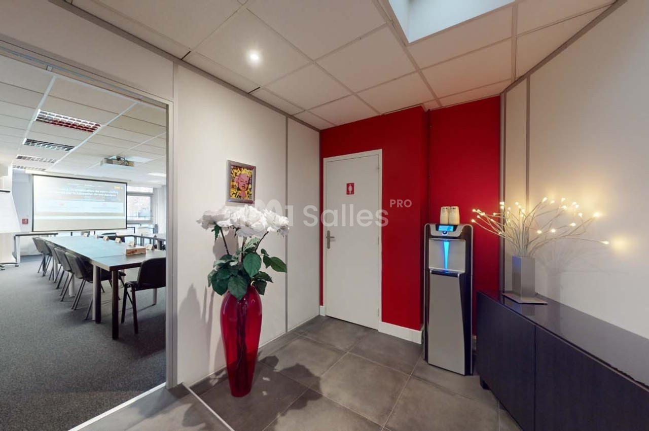 Vue d'un couloir avec une porte rouge, un distributeur d'eau, un vase de fleurs et une salle de réunion en arrière-plan.