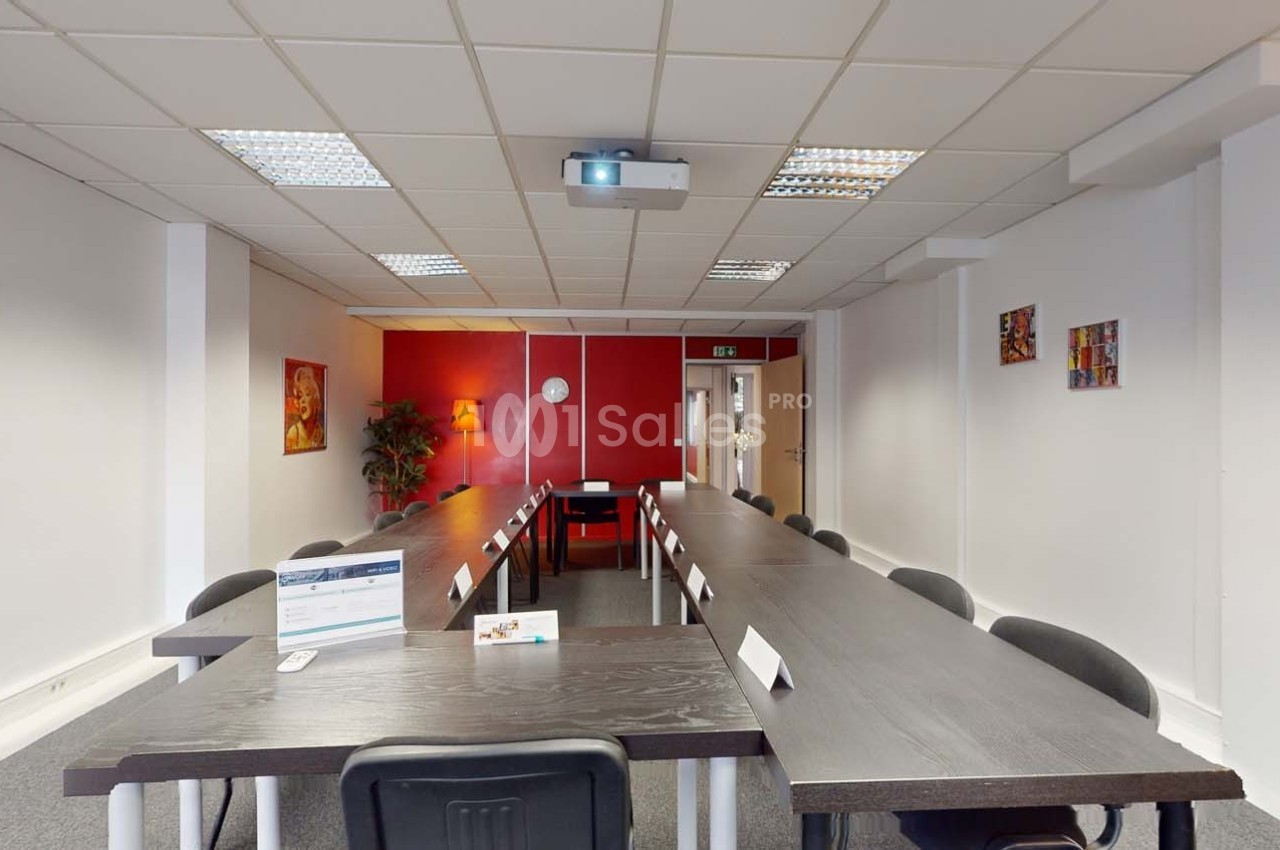 Salle de réunion lumineuse avec tables noires disposées en U, chaises noires et un projecteur fixé au plafond.