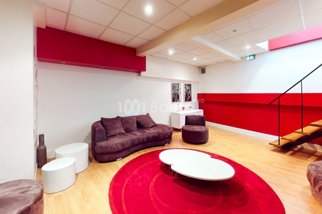Salon moderne avec canapé violet, tables basses blanches, tapis rouge rond et escalier en bois dans un espace lumineux.