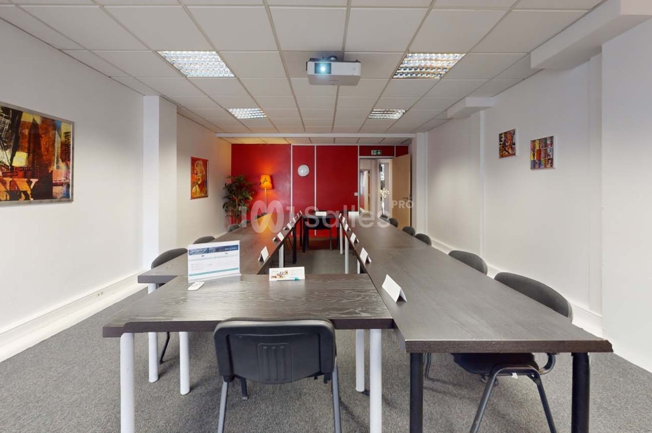 Salle de réunion lumineuse avec de longues tables noires disposées en U, chaises noires et projecteur au plafond.