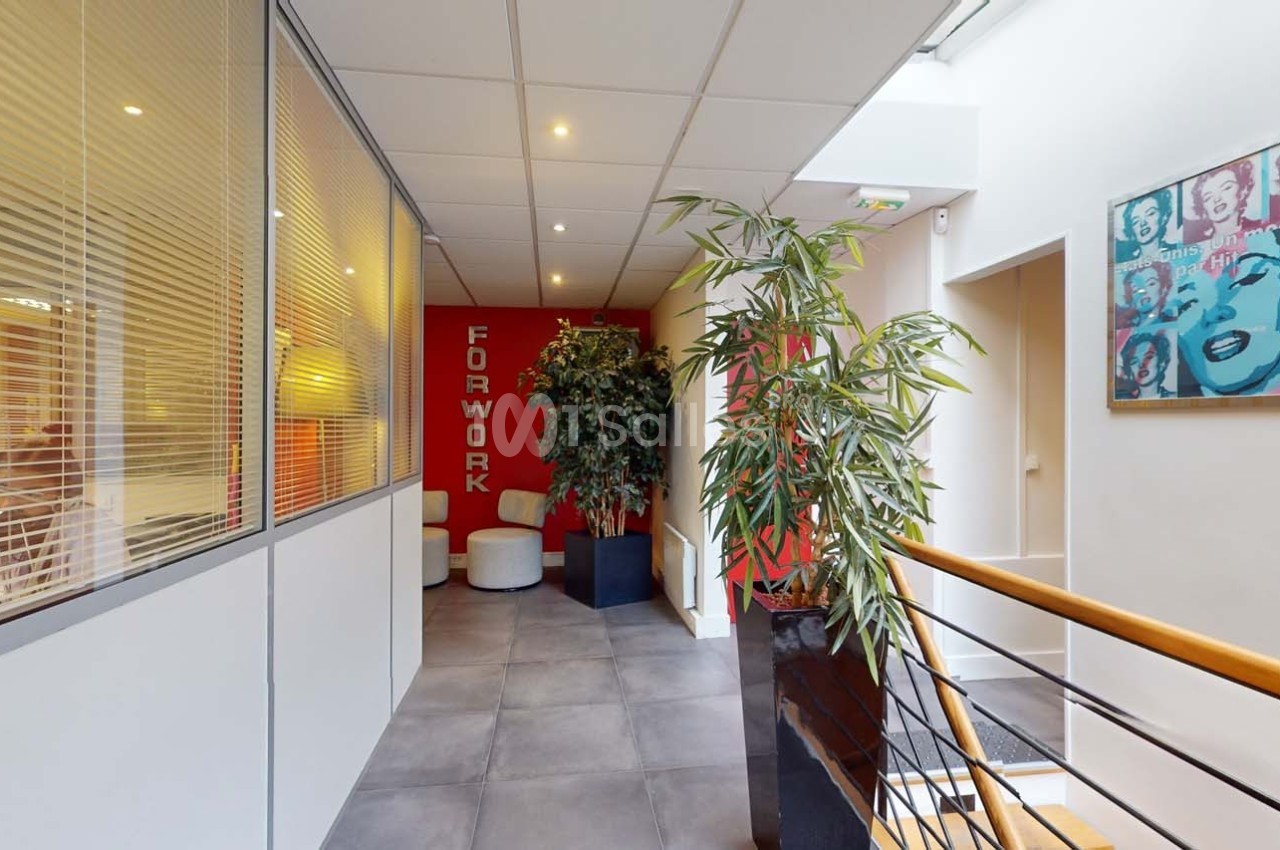 Couloir lumineux avec plantes, cloison vitrée à gauche, mur rouge avec inscription ’FOR WORK’ et tableau coloré à droite.