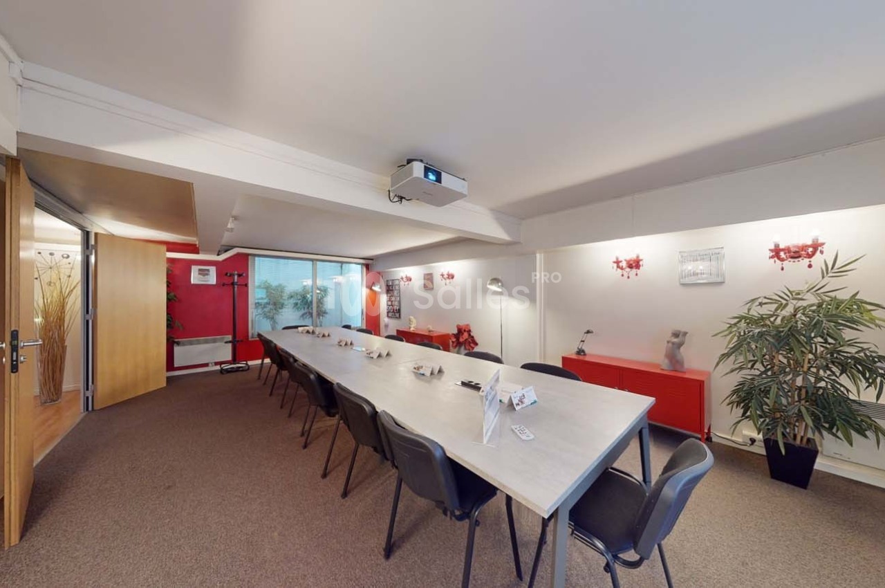 Salle de réunion lumineuse avec une grande table rectangulaire, des chaises noires et un projecteur au plafond.
