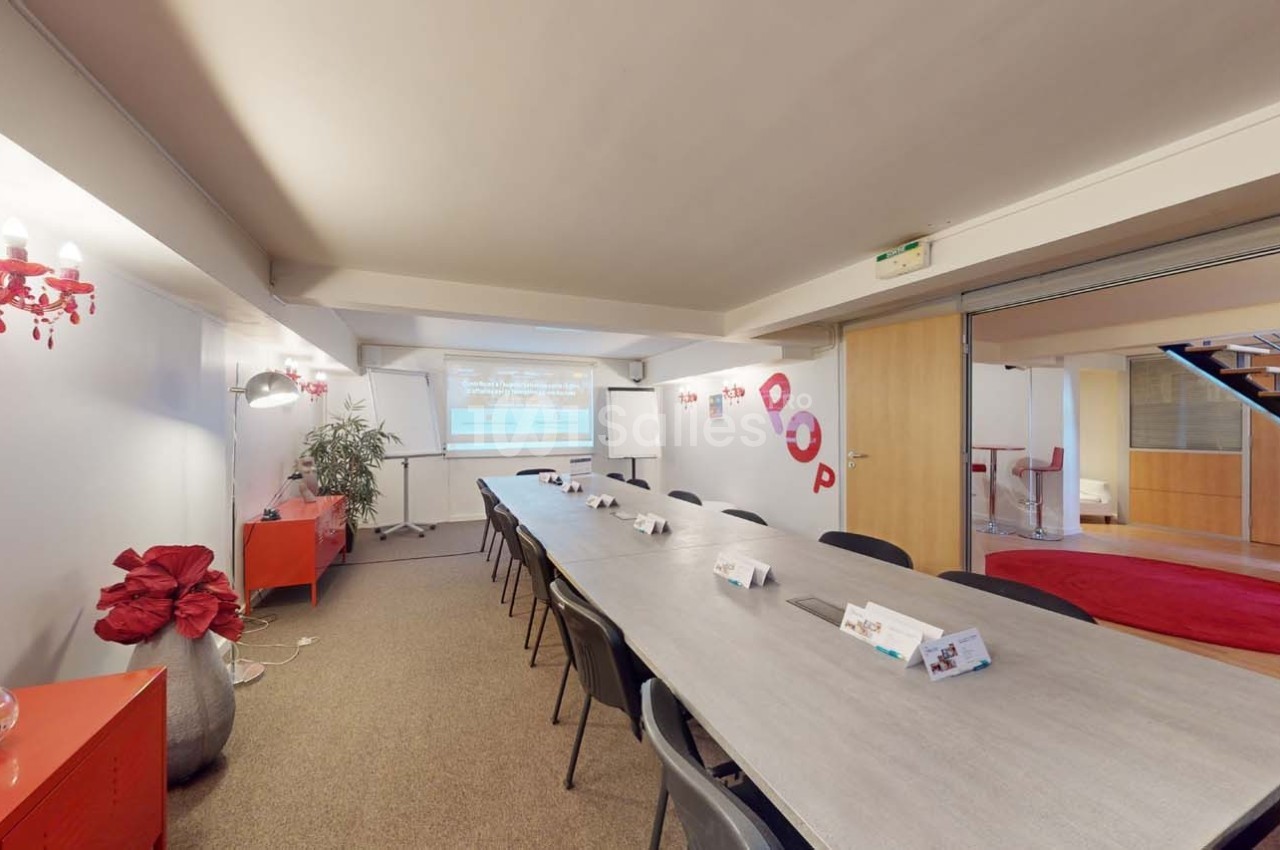 Salle de réunion lumineuse avec une grande table rectangulaire, chaises noires et écran de projection en fond.
