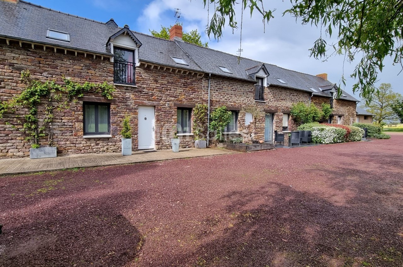 Façade d'une maison en pierre avec toit en ardoise, entourée de végétation et donnant sur une cour en gravier rouge.