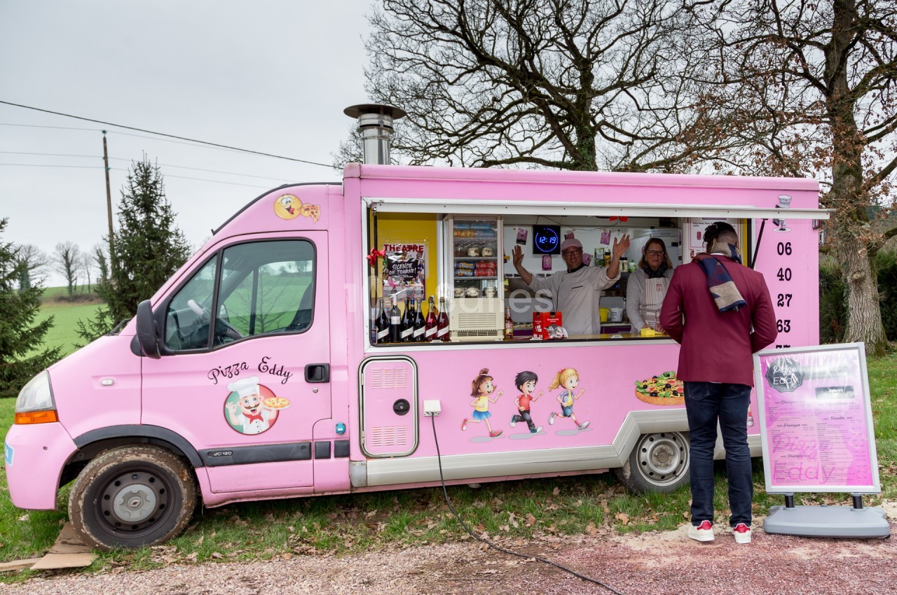 Camion-restaurant rose servant des pizzas, avec un client commandant devant et deux personnes à l'intérieur.