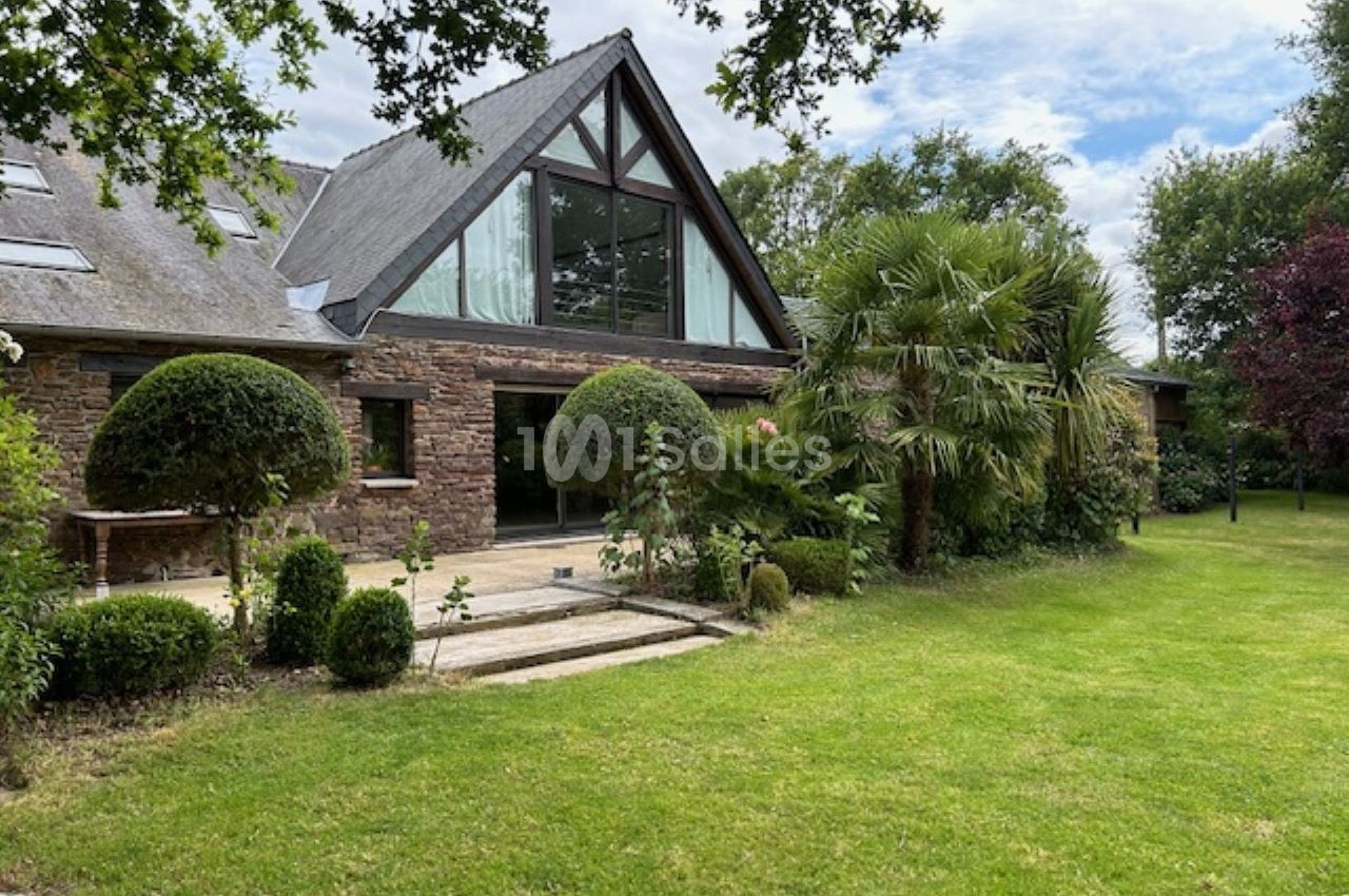 Maison en pierre avec grande baie vitrée, entourée d'un jardin arboré et d'une pelouse bien entretenue.