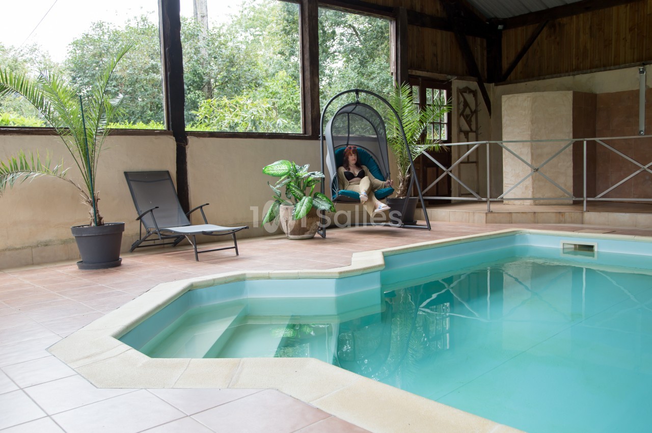 Piscine intérieure avec carrelage beige, transat, plantes en pot et une personne assise dans une chaise suspendue.