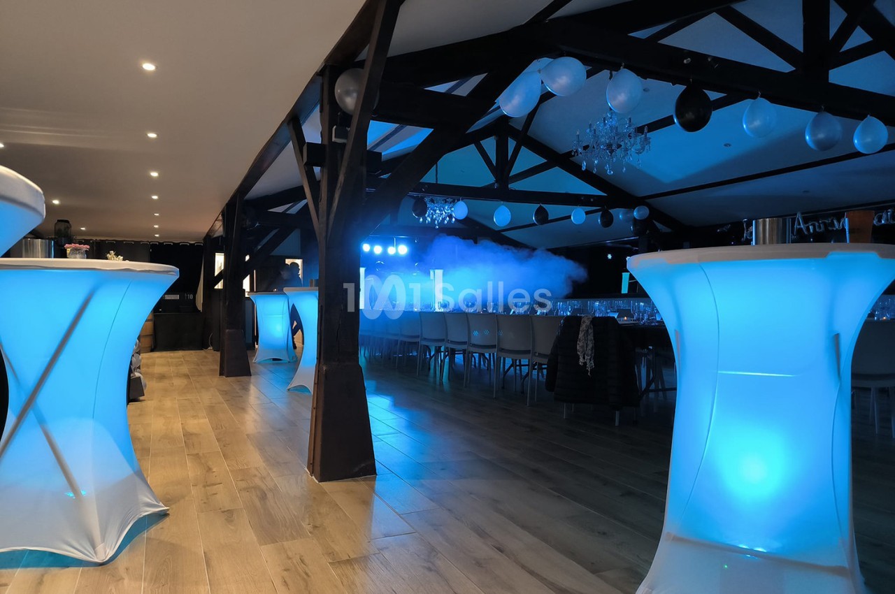 Salle de réception avec tables alignées, éclairage bleu et décorations suspendues sous une charpente en bois.