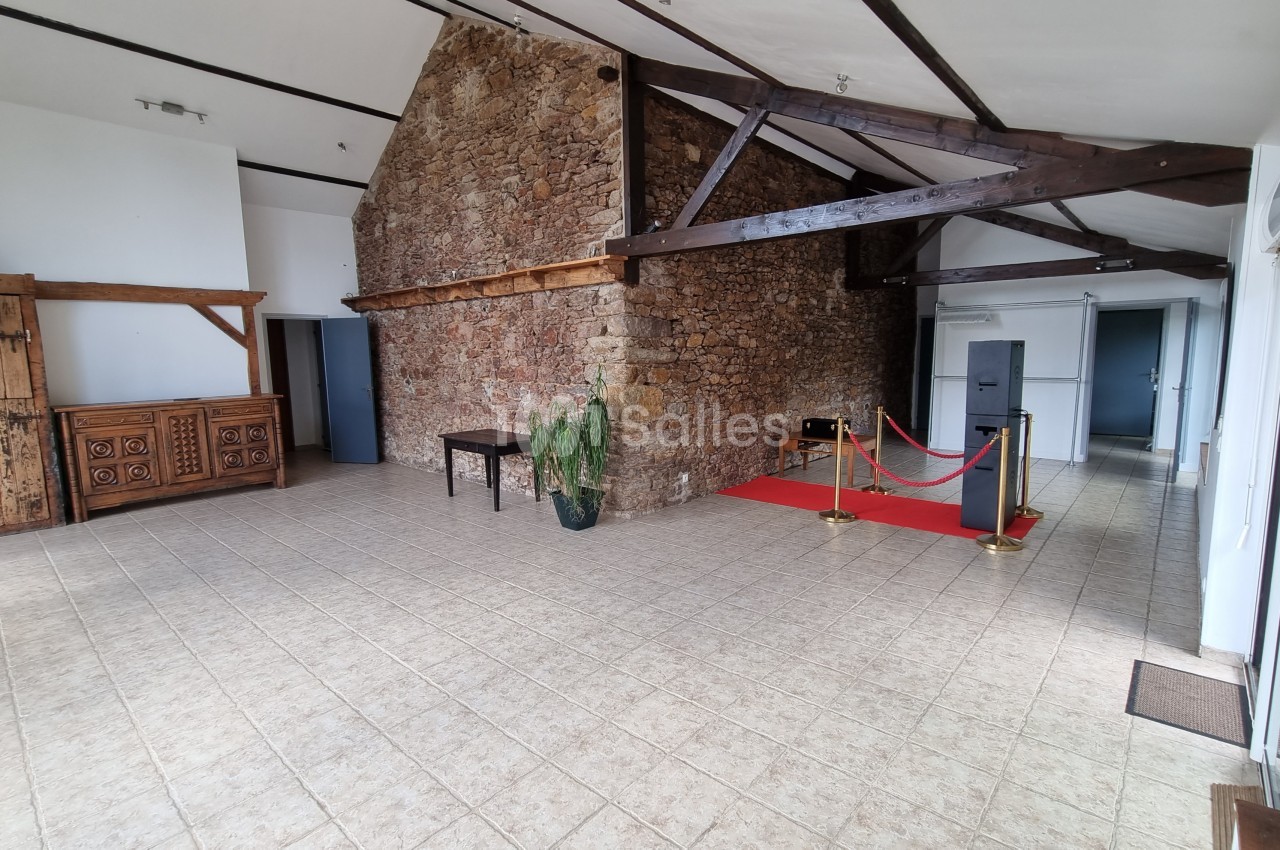 Salle lumineuse avec murs en pierre, poutres apparentes, carrelage beige, tapis rouge et mobilier en bois rustique.