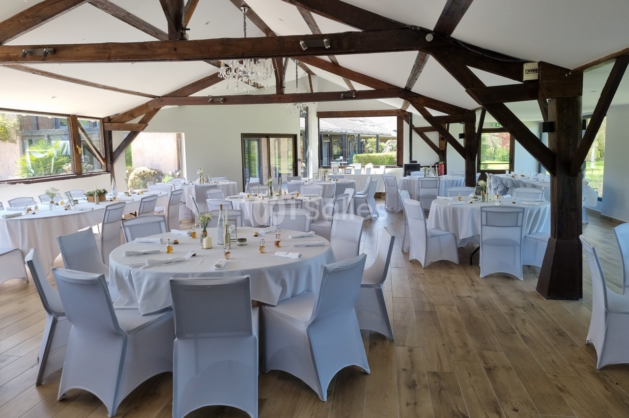 Salle de réception lumineuse avec tables rondes dressées, chaises blanches et poutres apparentes en bois.
