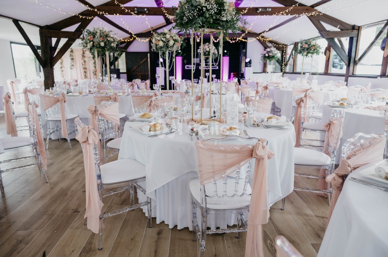 Salle de réception décorée pour un mariage avec des tables rondes, nappes blanches et chaises ornées de rubans roses.