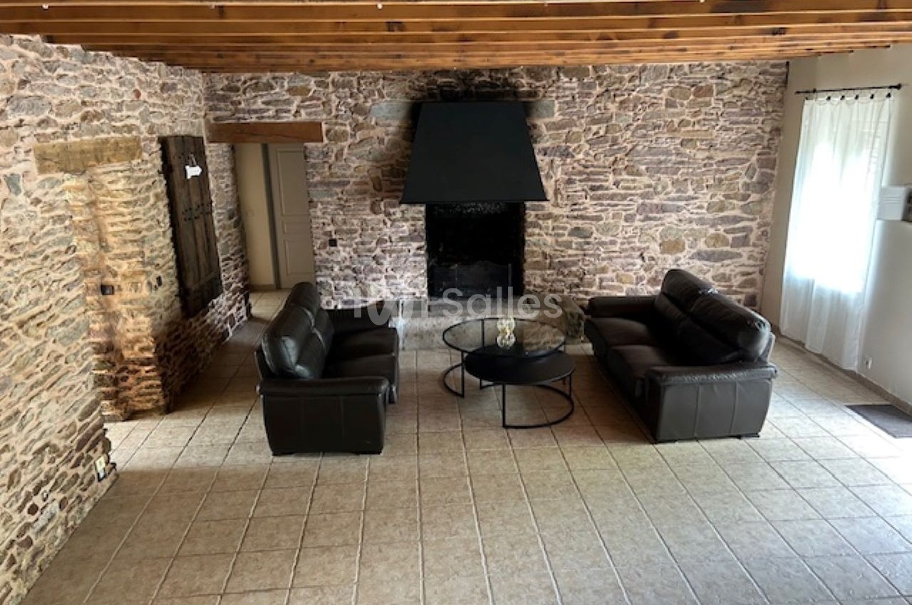 Salon avec murs en pierre apparente, plafond à poutres en bois, cheminée noire et mobilier en cuir brun.