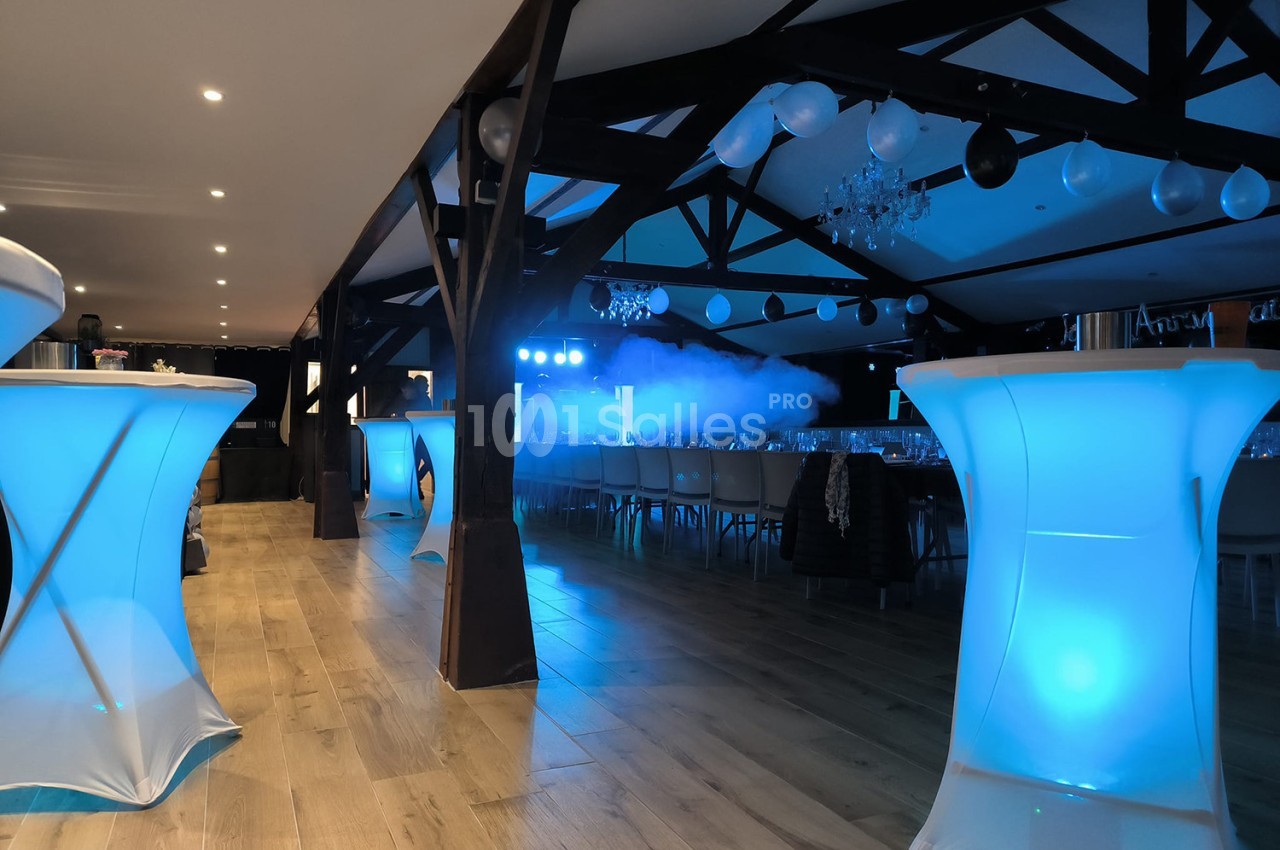 Salle de réception avec tables alignées, éclairage bleu et décorations suspendues sous une charpente en bois.