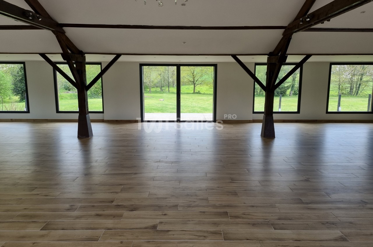 Salle vide avec parquet en bois, poutres apparentes et grandes baies vitrées donnant sur un espace vert.