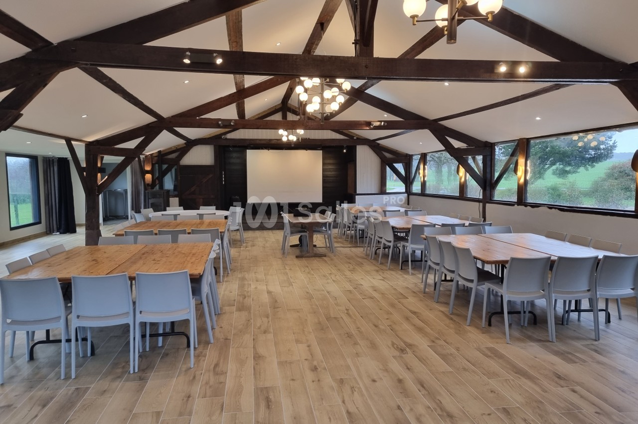 Salle spacieuse avec charpente en bois, tables et chaises disposées pour un événement, éclairage naturel et artificiel.