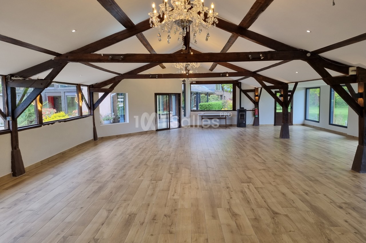 Salle spacieuse avec parquet en bois, poutres apparentes et lustre, entourée de grandes fenêtres donnant sur un jardin.