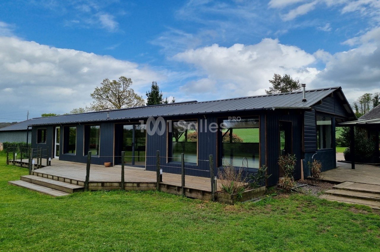 Maison moderne en bois sombre avec de grandes baies vitrées, entourée de pelouse et située dans un environnement verdoyant.