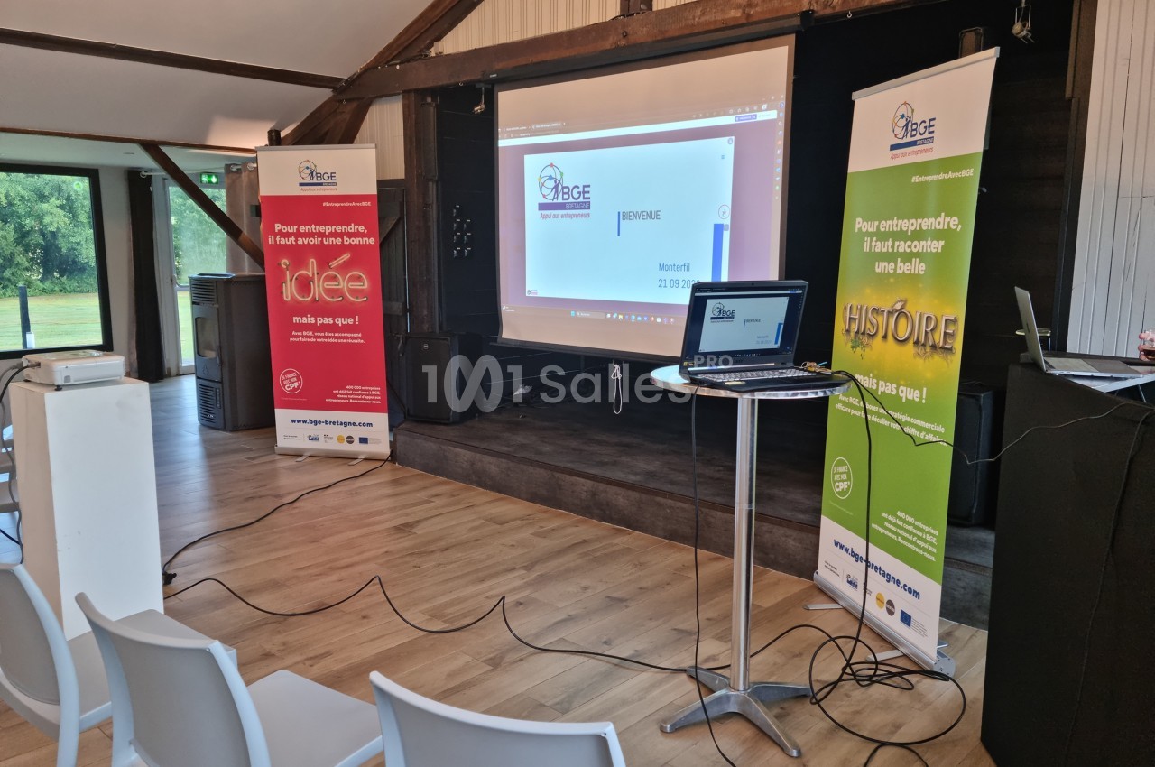 Salle de conférence avec écran de projection, ordinateur portable et affiches promotionnelles sur les côtés.