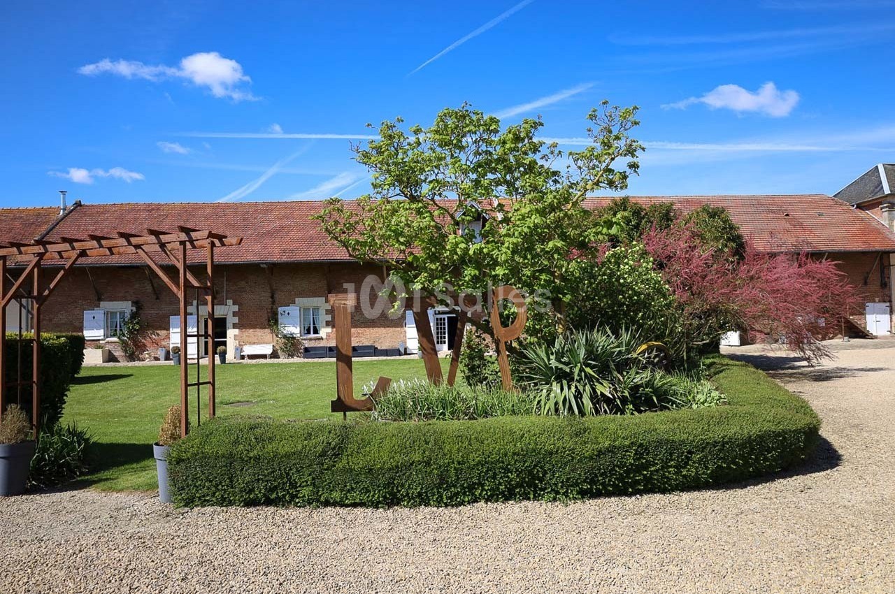 Jardin paysager avec arbustes taillés, sculptures métalliques et bâtiment en briques sous un ciel bleu.