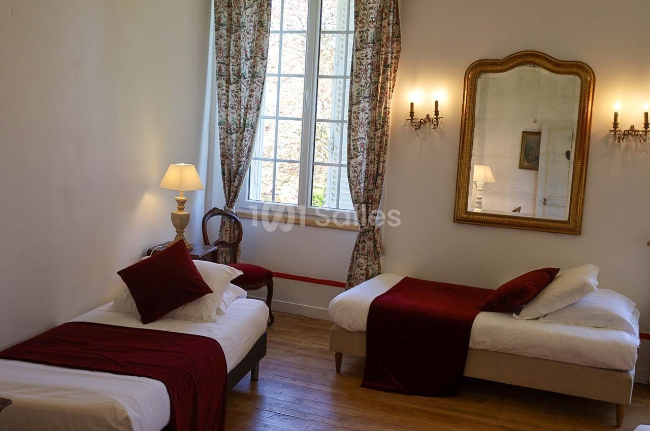 Chambre lumineuse avec deux lits simples, parquet en bois, grande fenêtre, miroir doré et rideaux fleuris.
