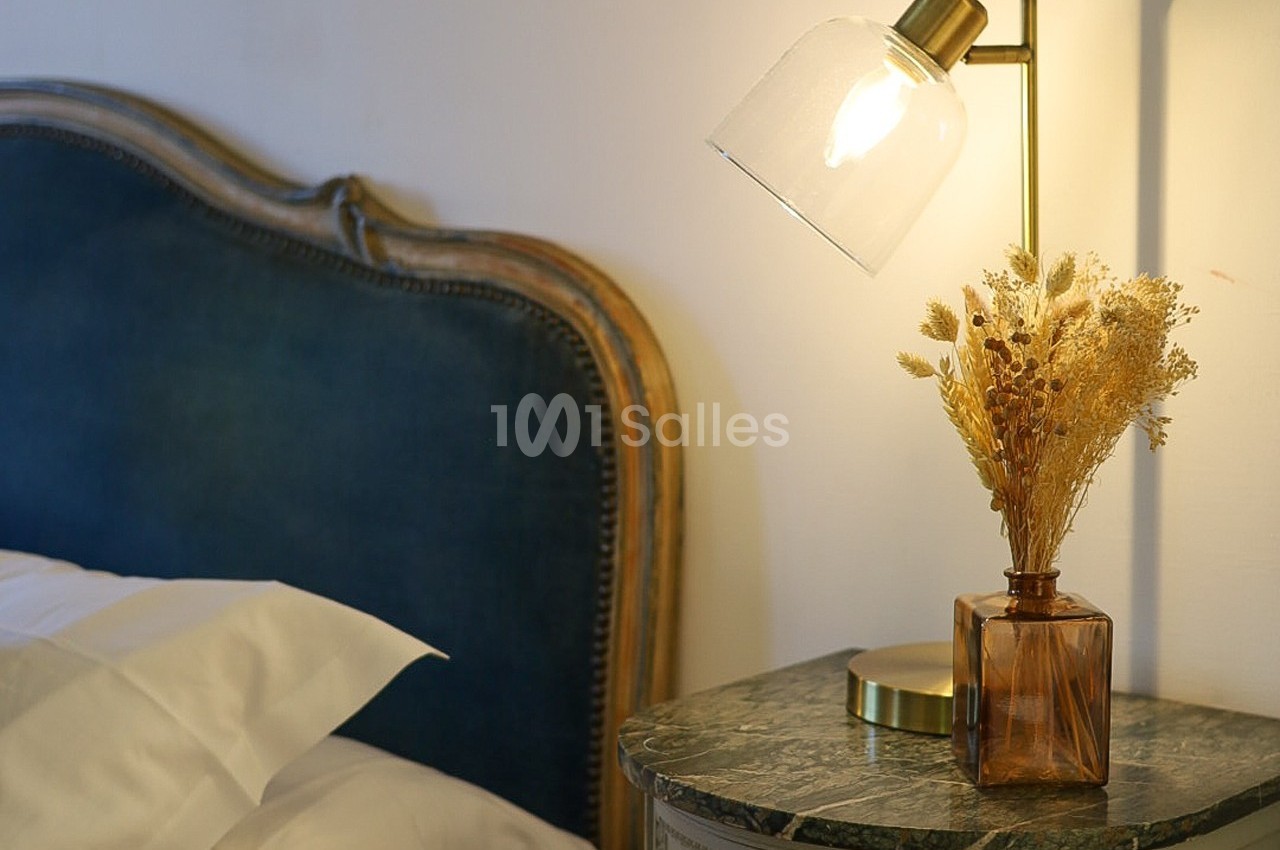 Lampe allumée sur une table de chevet en marbre avec un vase de fleurs séchées, près d'un lit avec tête de lit bleue.