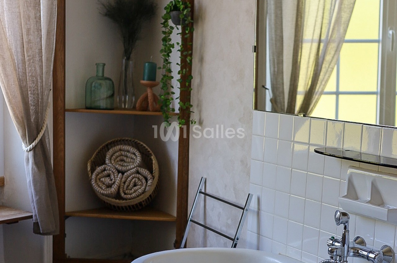 Salle de bain lumineuse avec baignoire, étagère en bois, serviettes roulées et rideaux laissant passer la lumière.