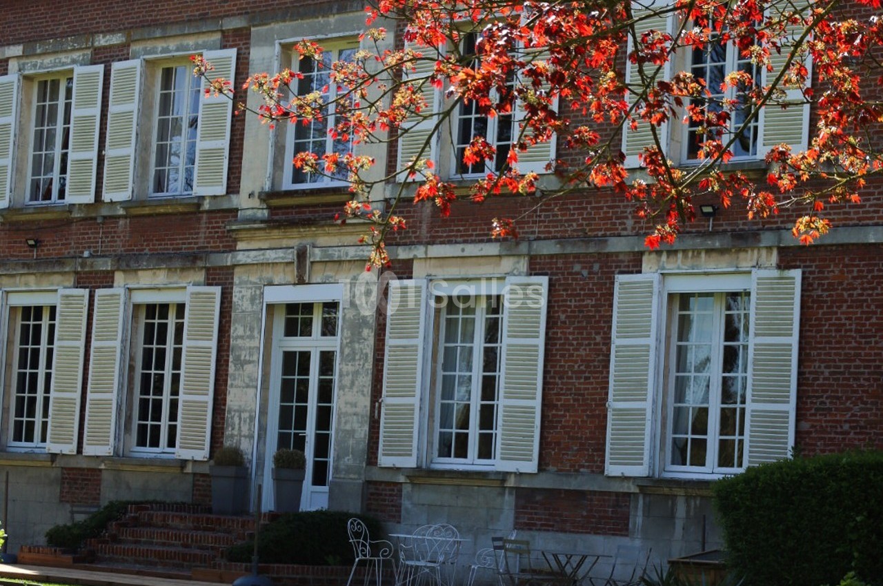 Façade d'une maison en briques rouges avec volets blancs, entourée d'un jardin et d'un arbre aux feuilles rouges.
