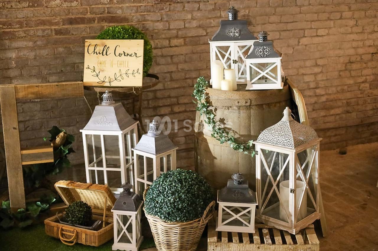 Lanternes blanches décoratives posées sur un tonneau et des paniers, entourées de plantes et bougies dans un décor rustique.