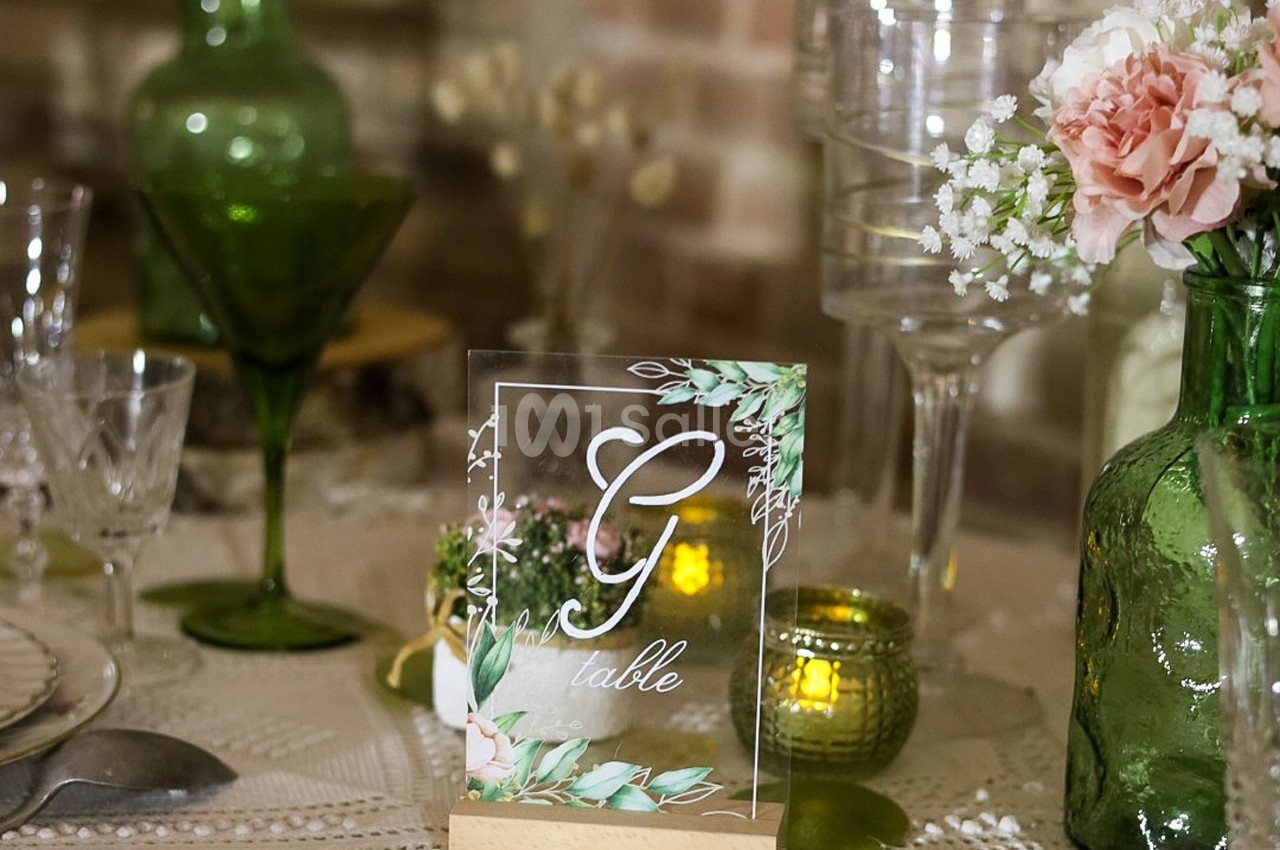 Carte de table décorative avec lettre ’G’, entourée de fleurs et verres sur une table élégamment dressée.