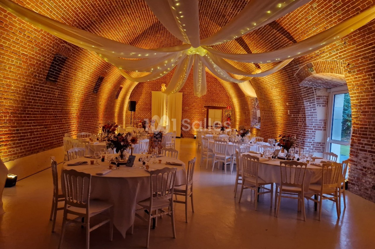 Salle voûtée en briques rouges décorée de guirlandes lumineuses et de tables rondes dressées pour un événement.