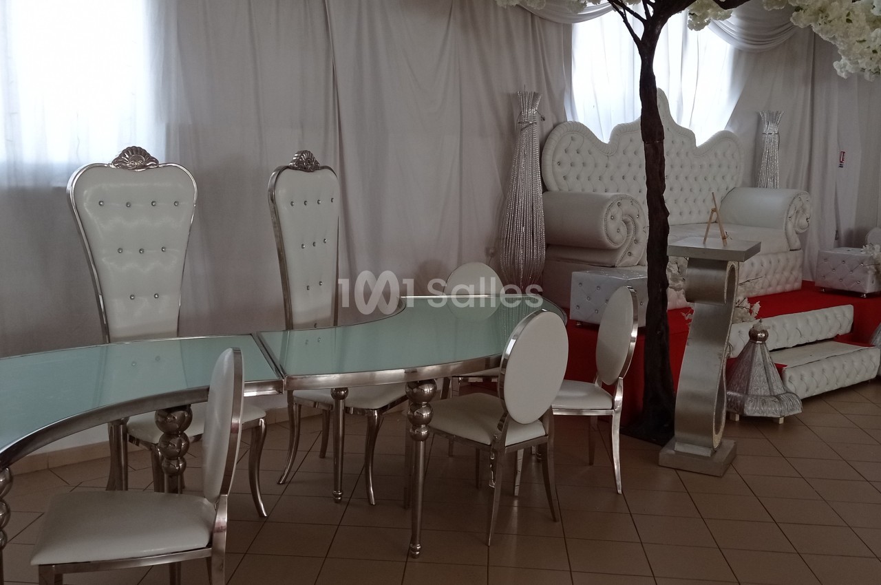 Salle de réception avec mobilier blanc et argenté, incluant des fauteuils capitonnés, une table en verre et des décorations…