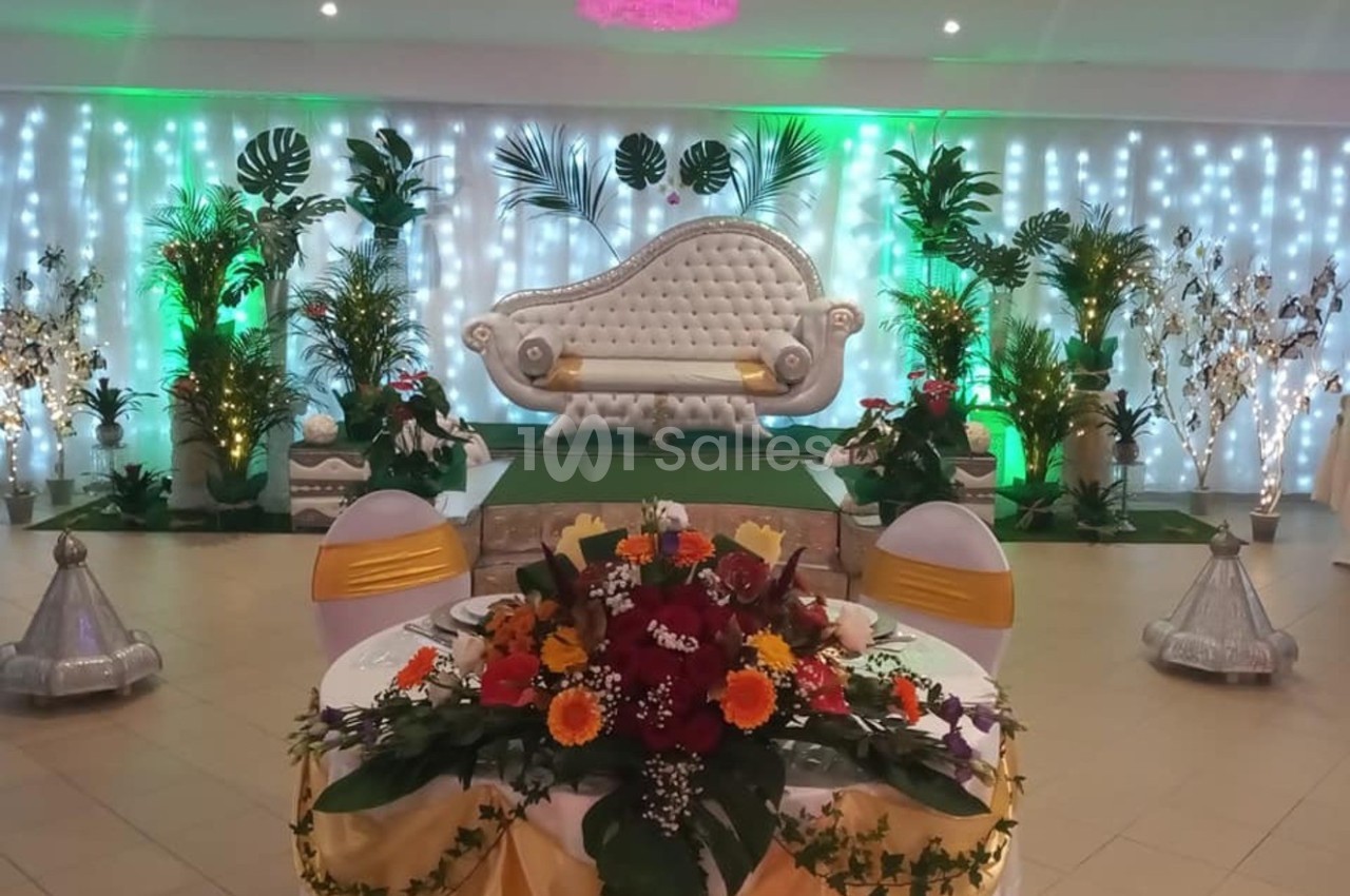 Salle décorée pour un événement avec un canapé élégant, des plantes, des lumières colorées et une table ornée de fleurs.