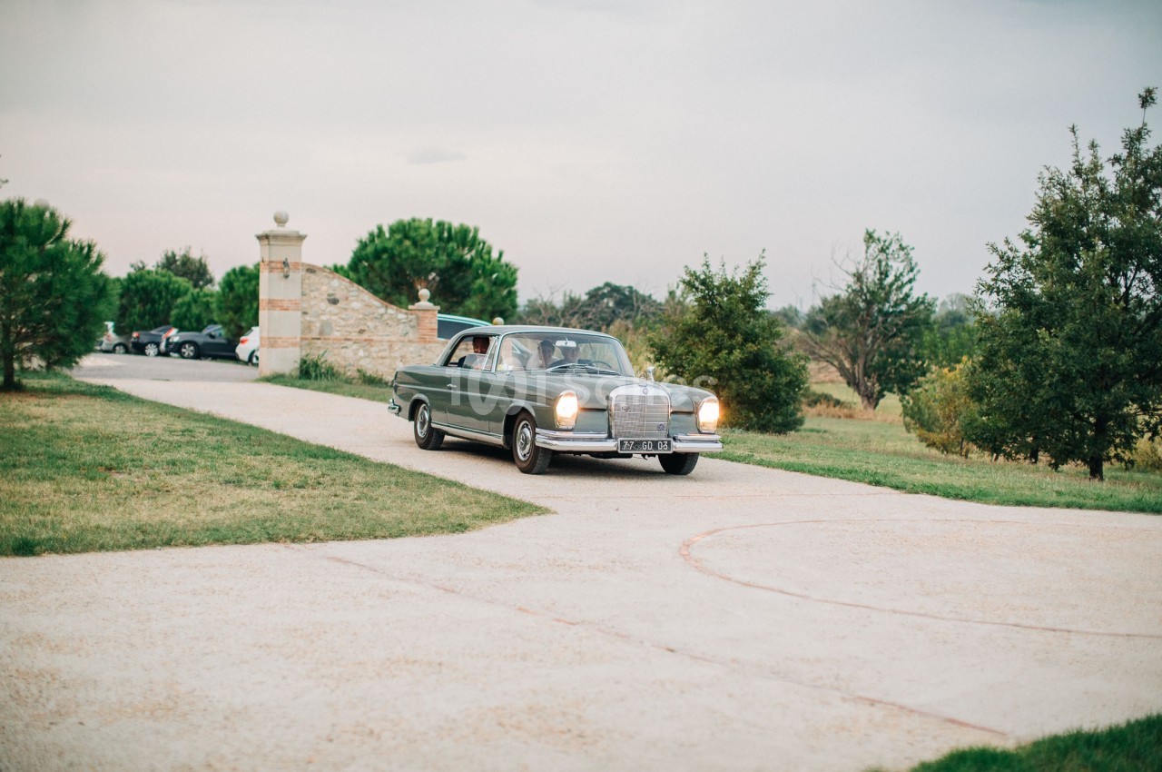Voiture ancienne gris métallisé roulant sur une allée bordée de pelouse, avec un portail en pierre à l'arrière-plan.