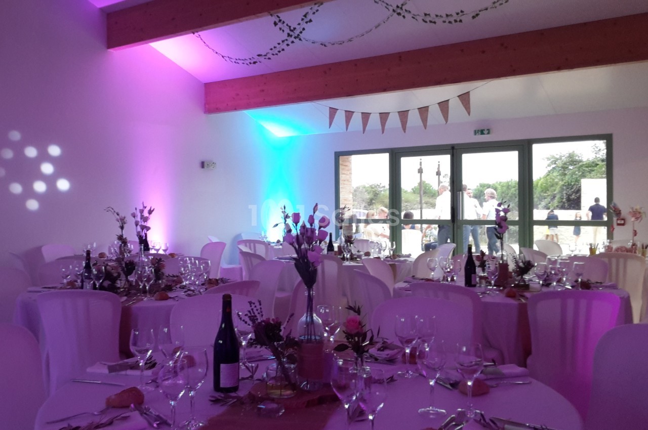 Salle de réception décorée pour un événement, avec tables dressées, nappes blanches et éclairage coloré.