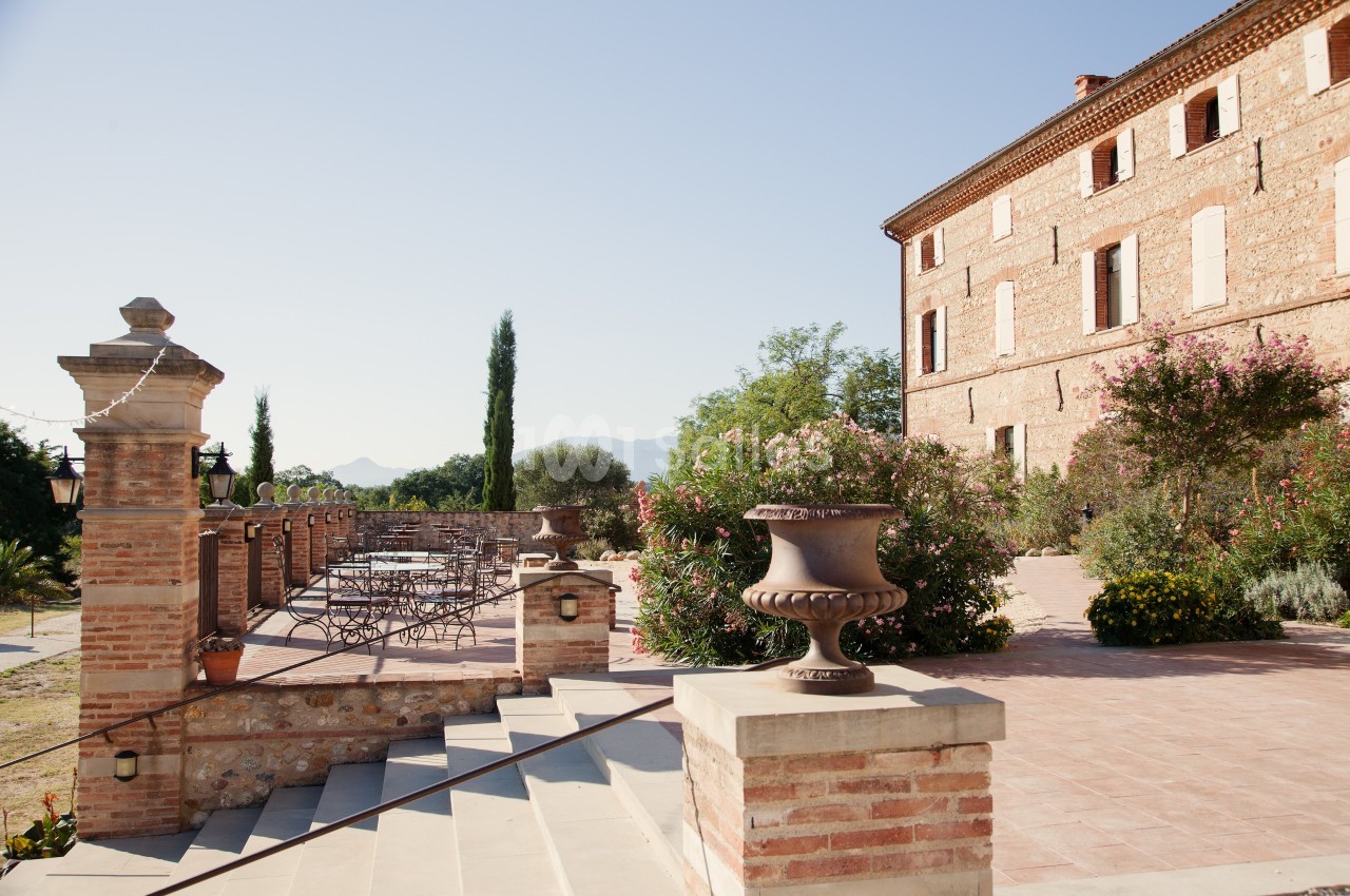 Terrasse ensoleillée avec tables et chaises, entourée de plantes, près d'un bâtiment en briques dans un cadre rural.