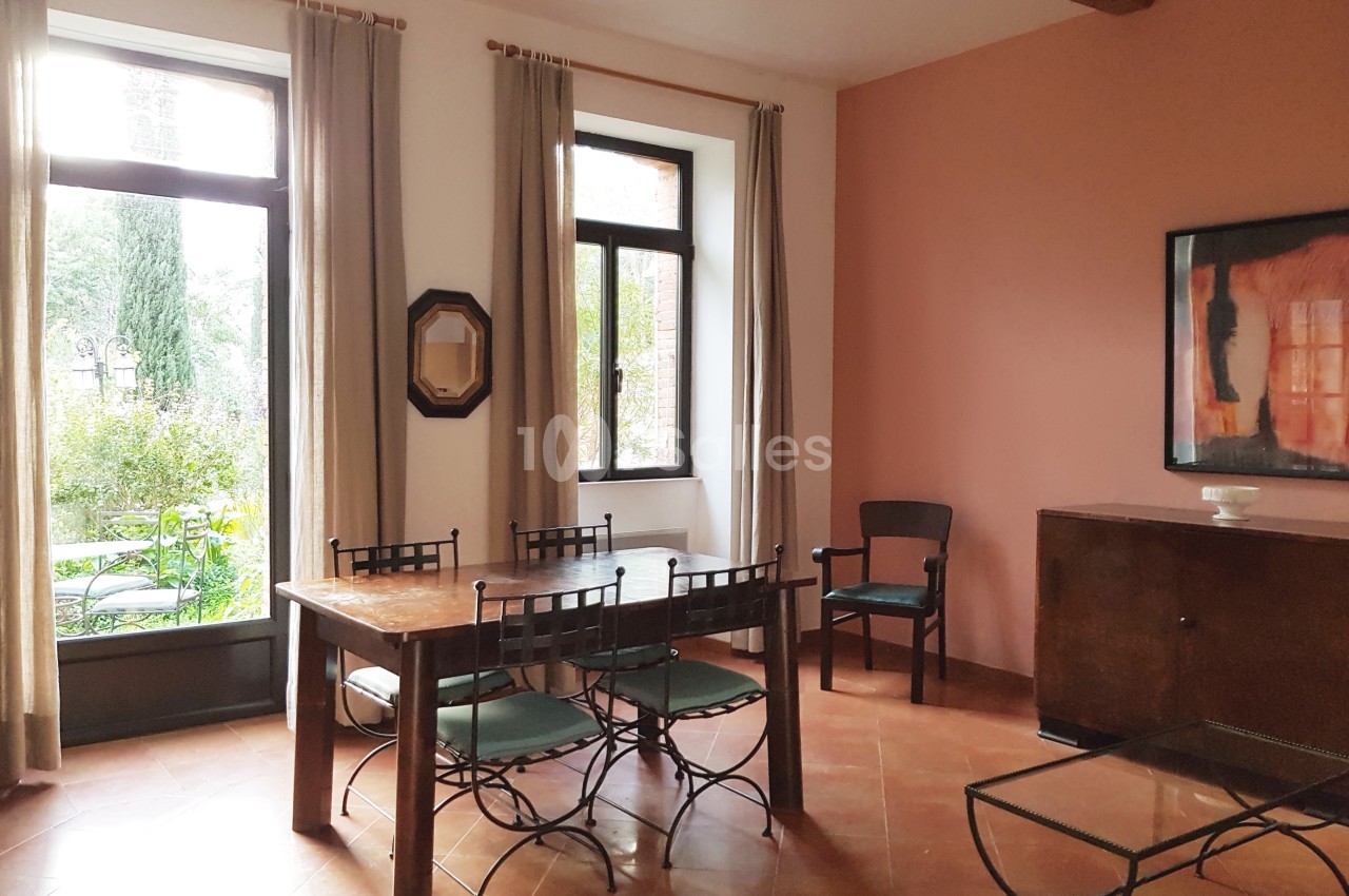 Salle à manger lumineuse avec table en bois, chaises en métal, murs roses, grandes fenêtres et vue sur un jardin.