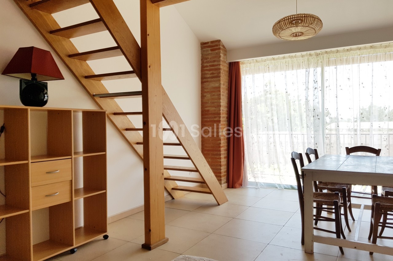 Escalier en bois dans un salon lumineux avec table, chaises, étagère et grande baie vitrée donnant sur l'extérieur.