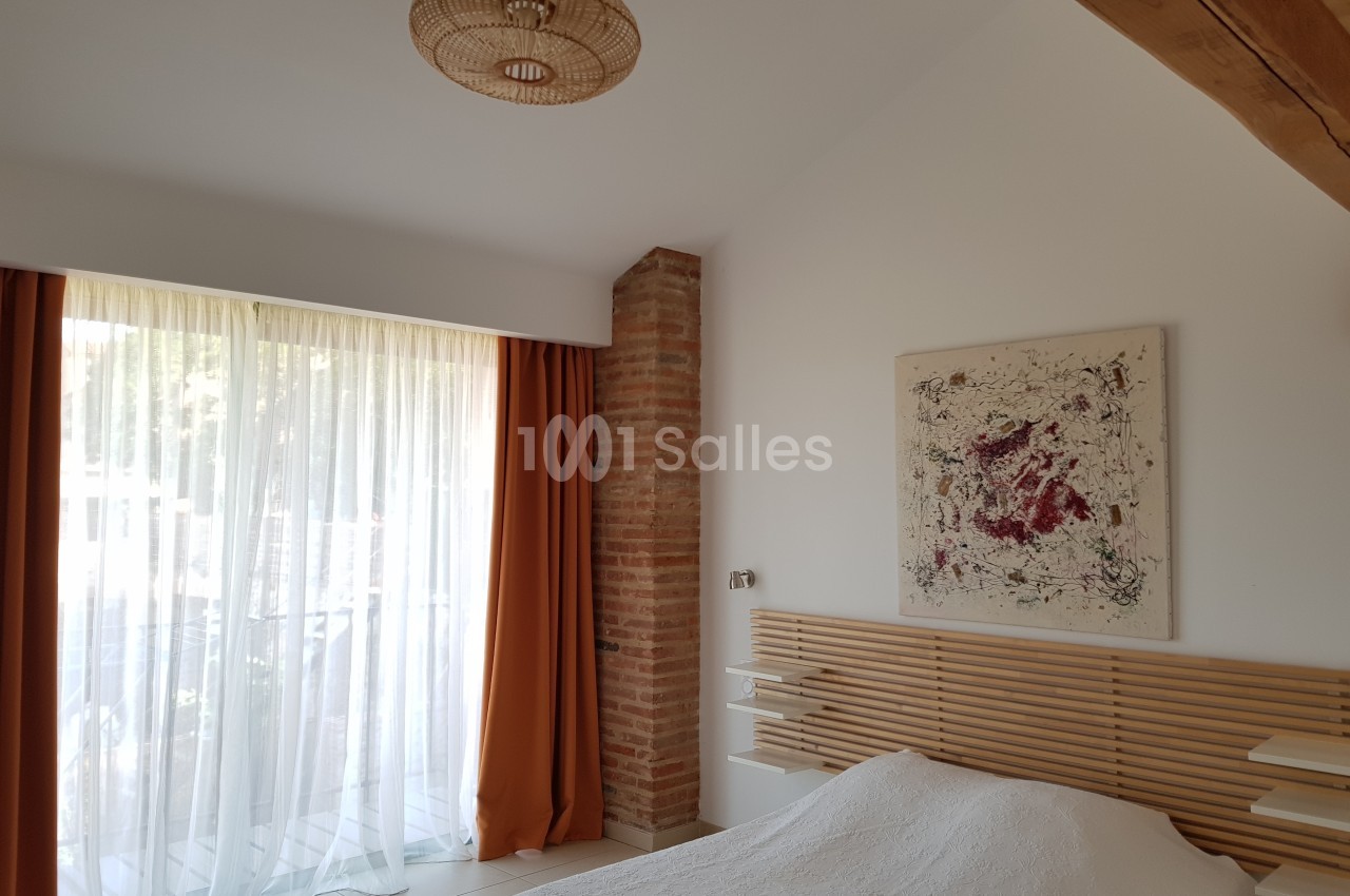 Chambre lumineuse avec lit, tête de lit en bois, tableau abstrait, rideaux orange et mur en briques apparentes.