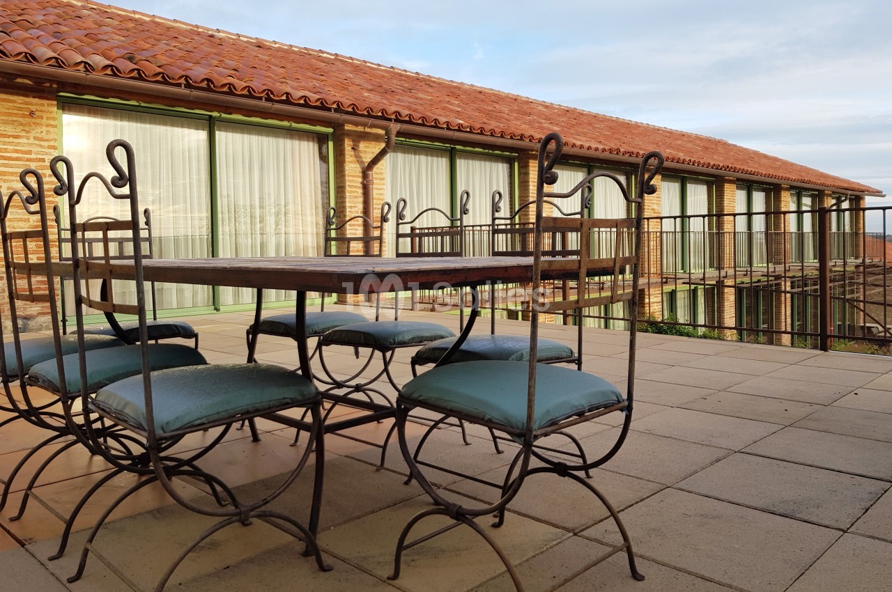 Table et chaises en fer forgé avec coussins bleus sur une terrasse ensoleillée devant un bâtiment en briques et grandes…