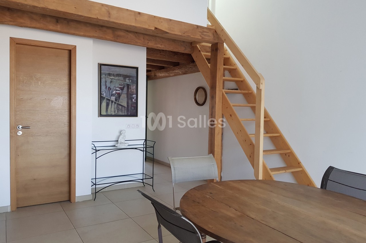 Intérieur lumineux avec table en bois, escalier en bois menant à une mezzanine, et porte en bois clair.