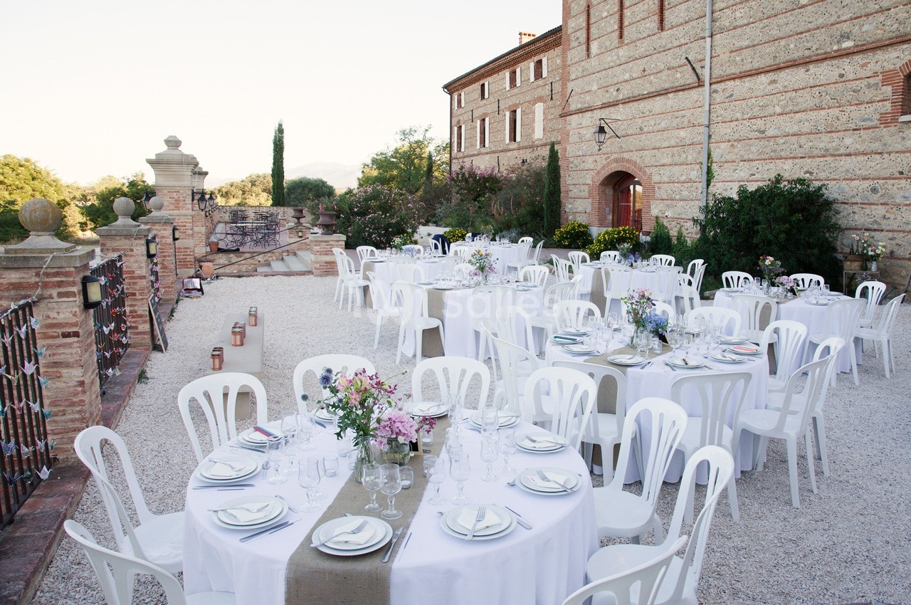 Tables rondes décorées pour un événement en plein air, disposées sur une terrasse en gravier près d'un bâtiment en briques.