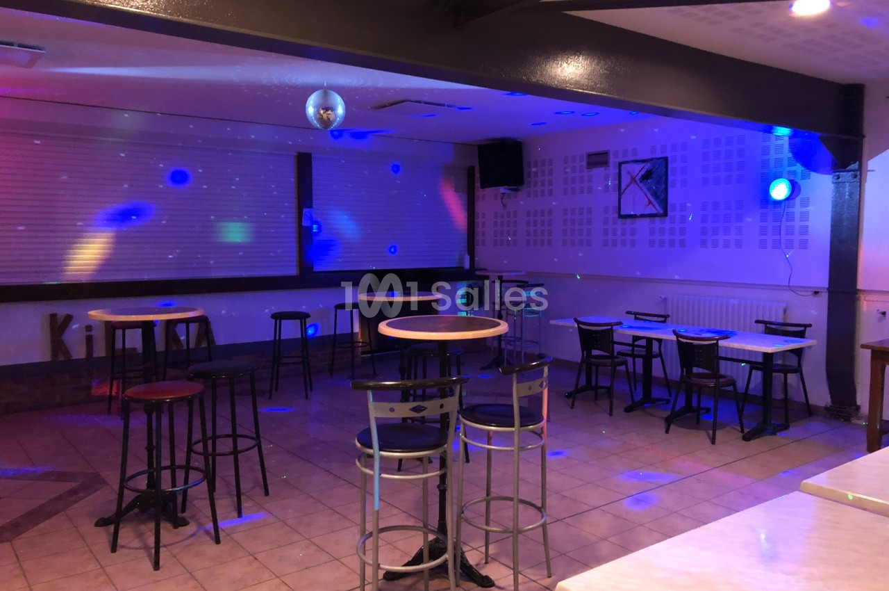 Salle vide avec tables hautes et basses, éclairage tamisé et spots colorés, ambiance de bar ou de petite discothèque.