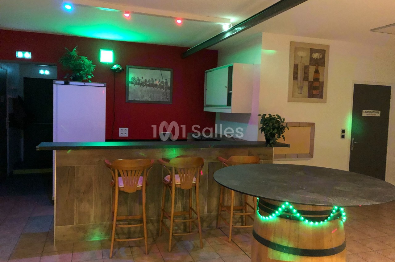 Bar avec comptoir en bois, tabourets, table ronde sur un tonneau, éclairage coloré et décoration murale.