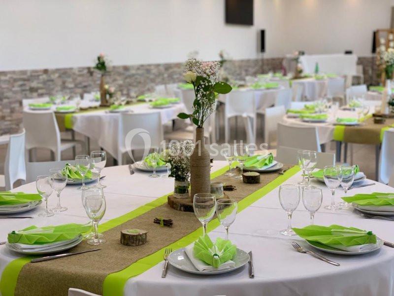 Salle décorée pour un événement, tables rondes avec nappes blanches, chemins de table verts et centres de table floraux.