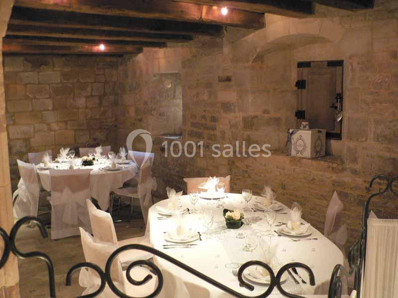 Salle en pierre avec poutres apparentes, tables rondes dressées pour un repas, décorée de nappes blanches et fleurs.
