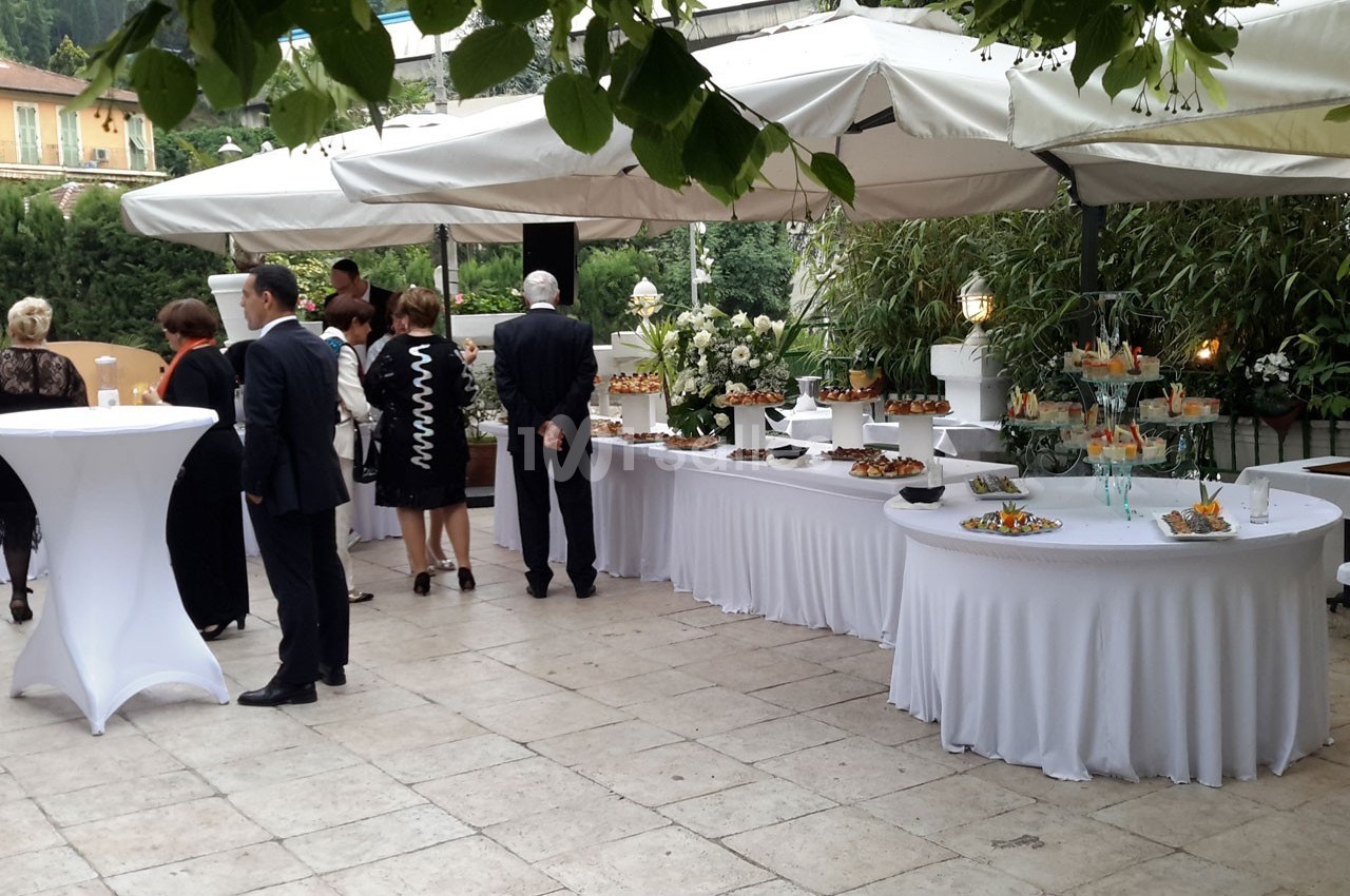 Buffet en extérieur avec tables garnies de plats, invités debout discutant sous des parasols blancs.