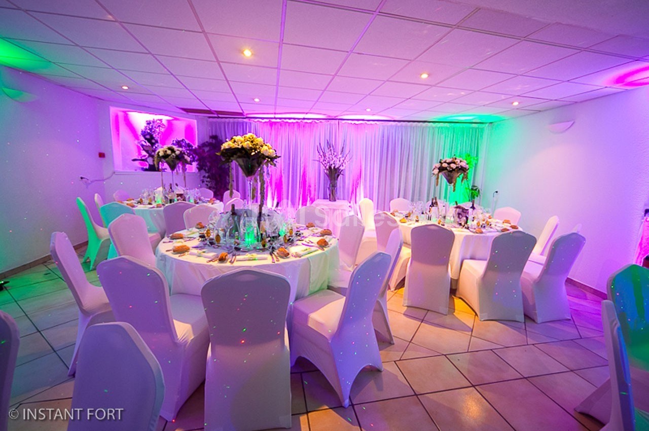Salle de réception décorée avec des tables rondes dressées, chaises blanches et éclairage coloré.