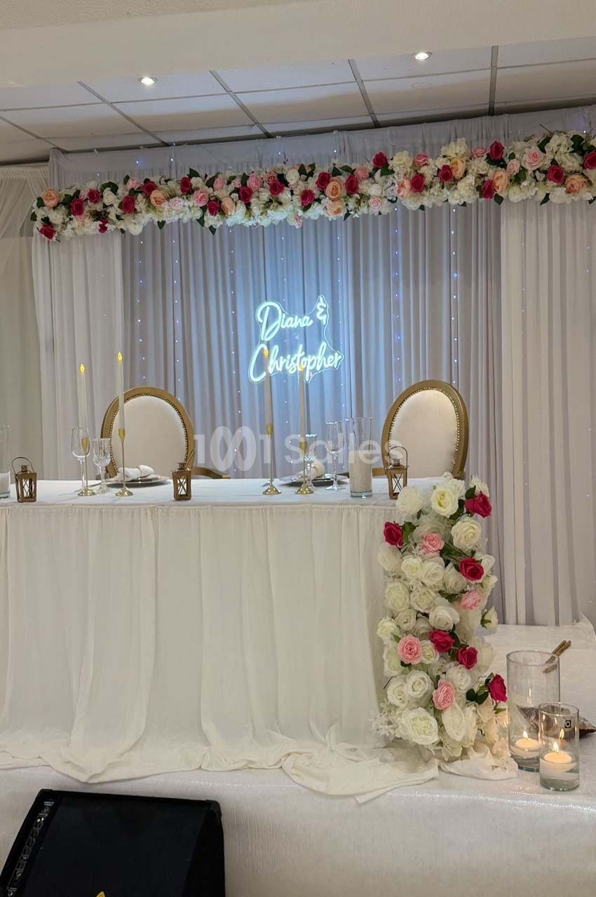 Table d'honneur décorée de fleurs blanches et roses, avec un panneau lumineux affichant des prénoms en arrière-plan.