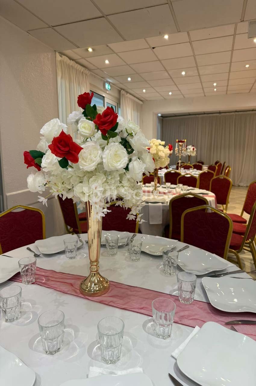 Salle de réception décorée avec des tables dressées, ornées de centres de table composés de roses rouges et blanches.